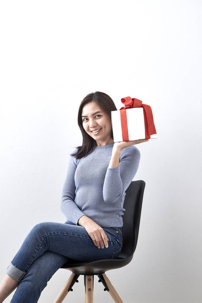 Kaunis seksikäs nuori aasialainen nainen lahjapakkaus istuu tuolilla valkoisella taustalla, kuva sisältää kopiotilaa
 - Valokuva, kuva