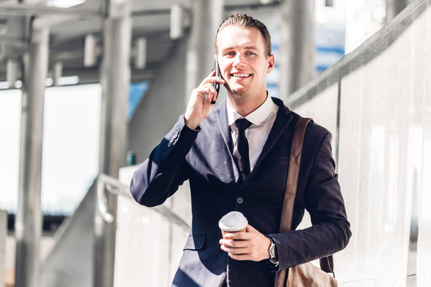 Porträt eines gutaussehenden Geschäftsmannes im schwarzen Anzug, der im Freien steht und sein Smartphone vor dem Hintergrund der modernen Großstadt nutzt. - Foto, Bild