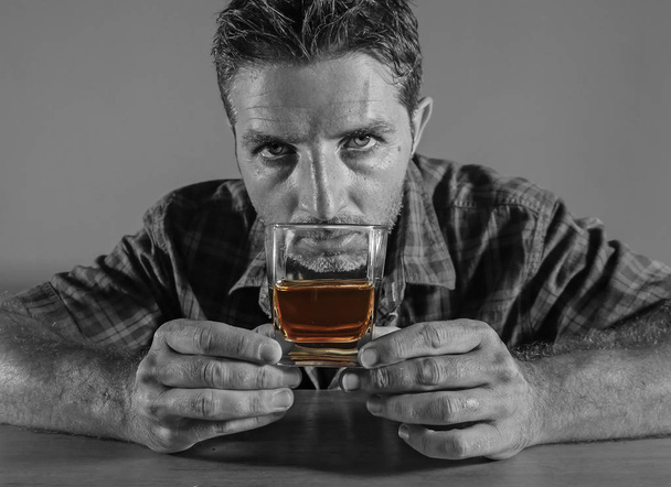 isolated black and white portrait of young messy depressed alcoholic man having a drink looking at whiskey glass feeling temptation of alcohol abuse in addiction and alcoholism problem - Foto, Imagen
