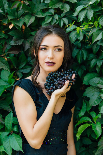 Portrait of young sexy brunette in black dress with black grapes. . - Foto, immagini
