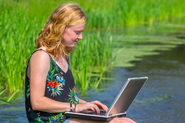 Fiatal vöröshajú nő dolgozik laptop közelében víz a természetben - Fotó, kép