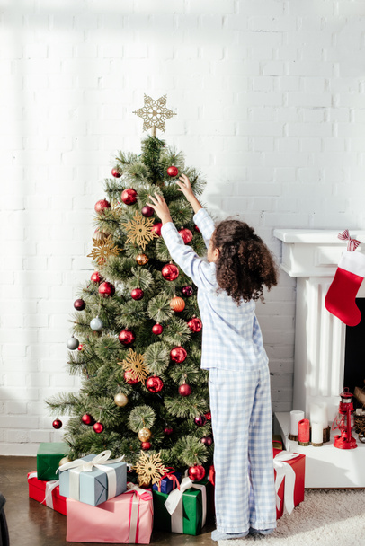 back view of african american child in pajamas decorating christmas tree with baubles at home - Foto, imagen