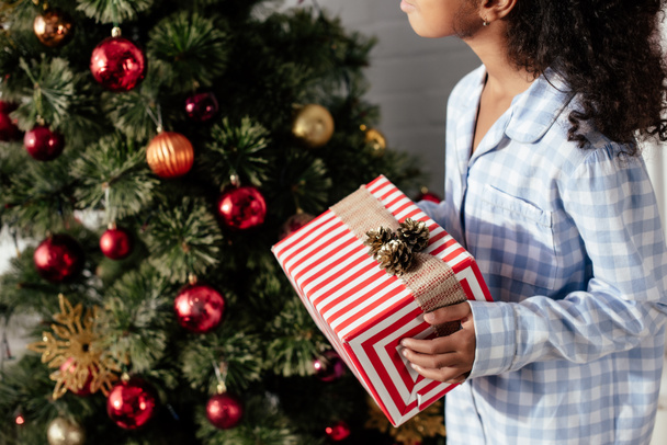 обрезанное изображение африканского американского ребенка в пижаме с рождественской подарочной коробкой дома
 - Фото, изображение