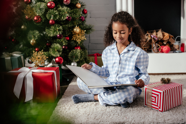cheerful adorable african american child in pajamas reading book near christmas tree at home - Valokuva, kuva
