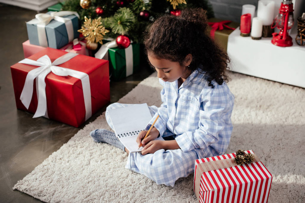 прелестный африканский американец в пижаме пишет что-то в тетрадь дома, рождественская концепция
 - Фото, изображение