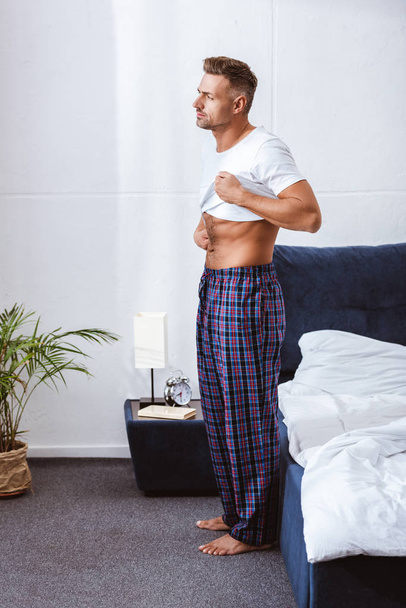 adult man putting on white t-shirt near bed at home - Foto, afbeelding