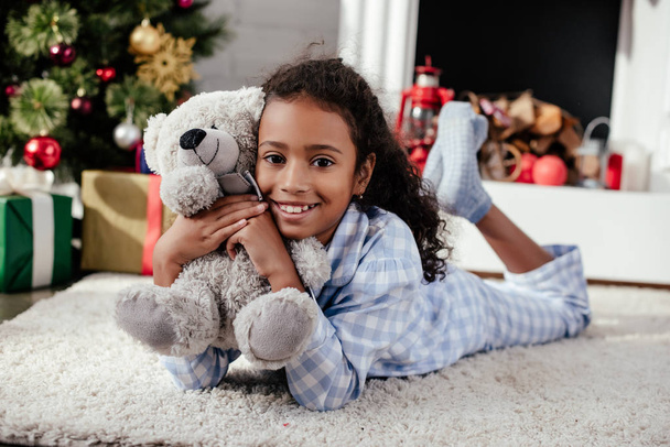 mosolygó imádnivaló afro-amerikai gyermek feküdt a padlón a teddy bear otthon, karácsonyi concept pizsamában - Fotó, kép