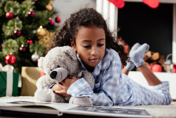 adorable african american child in pajamas with teddy bear looking at photo album on floor at home - Photo, Image