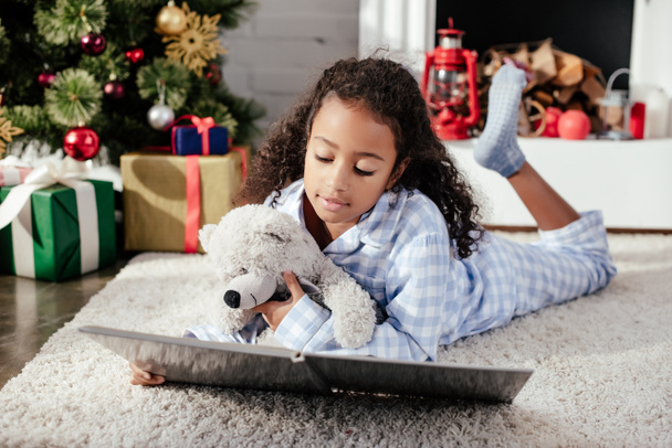 adorable african american child in pajamas with teddy bear reading book on floor at home, christmas concept - Fotografie, Obrázek