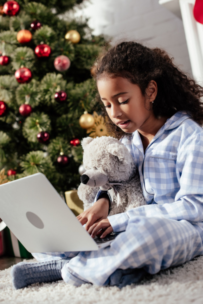 adorable african american child in pajamas with teddy bear using laptop at home, christmas concept - Foto, immagini