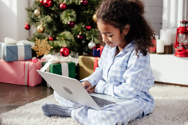 adorable african american child in pajamas using laptop at home, christmas concept - Photo, Image