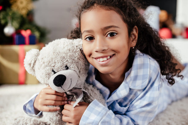 portrét rozkošný šťastné africké americké dítě v pyžamu s plyšovým medvědem při pohledu na fotoaparát doma - Fotografie, Obrázek