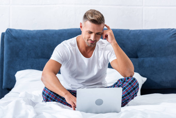 красивый мужчина фрилансер сидит на кровати и использует ноутбук дома
 - Фото, изображение