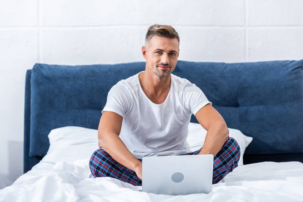 sourire freelance masculin regardant la caméra et assis sur le lit avec ordinateur portable à la maison
 - Photo, image