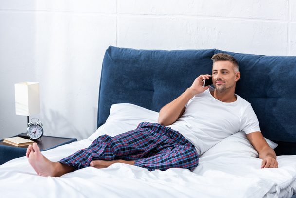 весёлый взрослый мужчина разговаривает по смартфону, лежа дома в постели
 - Фото, изображение