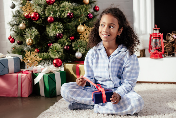 happy adorable african american child in pajamas opening christmas gift at home and looking away - Foto, imagen