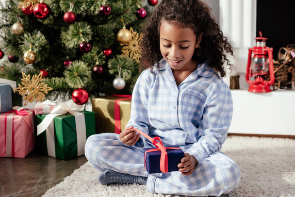 excited adorable african american child in pajamas opening christmas gift at home - Fotografie, Obrázek