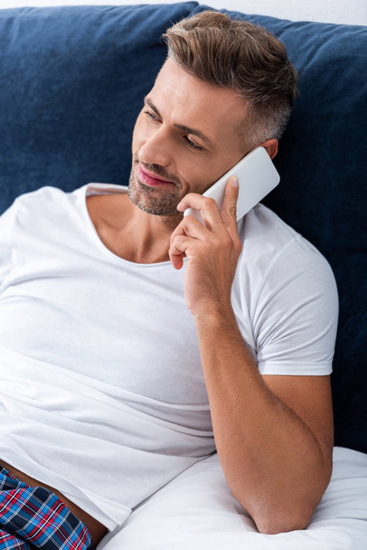 hombre adulto feliz hablando en el teléfono inteligente mientras está acostado en la cama en casa
 - Foto, Imagen