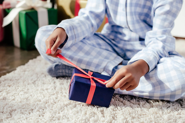 cropped image of african american child in pajamas opening christmas present on carpet at home - Foto, imagen
