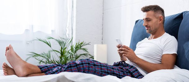 happy man in white t-shirt using smartphone during morning time in bed at home - Photo, image