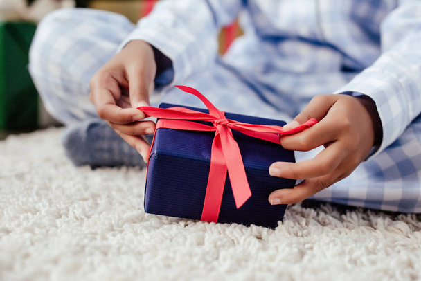 bijgesneden afbeelding van Afro-Amerikaanse kind in pyjama's openen de gift van Kerstmis op verdieping thuis - Foto, afbeelding