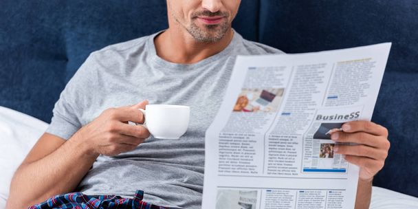 partial view of businessman with cup of coffee reading business newspaper in bed at home - Фото, изображение