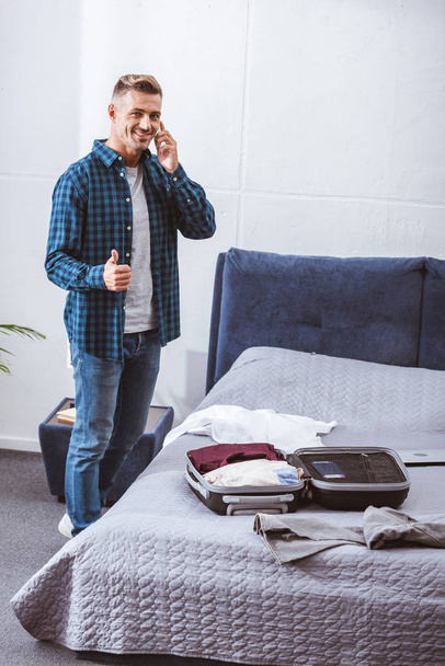 hymyilevä aikuinen mies puhuu älypuhelimella ja tekee peukalo ylös ele lähellä matkatavarat kotona
 - Valokuva, kuva