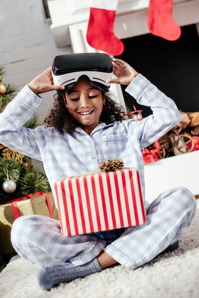 happy adorable african american child in pajamas and virtual reality headset looking at christmas gift at home - Photo, Image