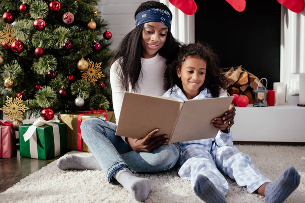 cheerful african american mother and daughter reading book together at home, christmas concept - Foto, afbeelding