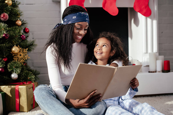 happy african american mother and daughter reading book together at home, christmas concept - Photo, Image