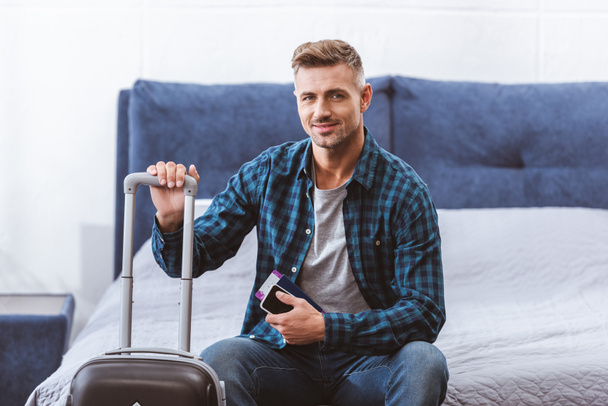szczęśliwy podróżnik mężczyzna siedzi na łóżku z smartphone i lot bilet w pobliżu kołowych worek w domu - Zdjęcie, obraz