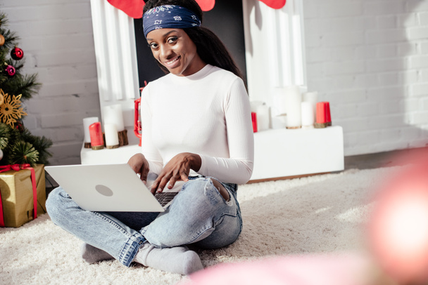 ελκυστική αφρικανική αμερικανική γυναίκα που χρησιμοποιούν φορητό υπολογιστή στο πάτωμα στο σπίτι και βλέπουν φωτογραφική μηχανή, Χριστούγεννα έννοια - Φωτογραφία, εικόνα