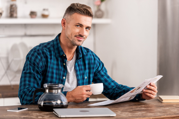 felice uomo con caffè tazza lettura giornale a tavola con computer portatile in cucina a casa
 - Foto, immagini