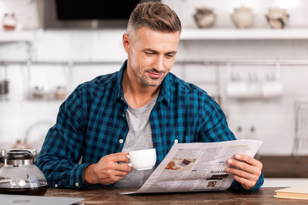 Glimlachende man in geruite hemd houden van kopje koffie en het lezen van de krant thuis - Foto, afbeelding