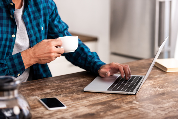 cropped shot of man holding cup of coffee and using laptop at home - Фото, зображення