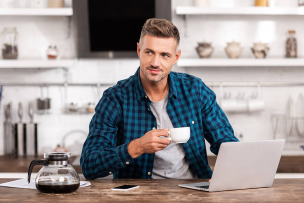 guapo sonriente hombre sosteniendo la taza de café y mirando hacia otro lado mientras usa el ordenador portátil en casa
 - Foto, imagen