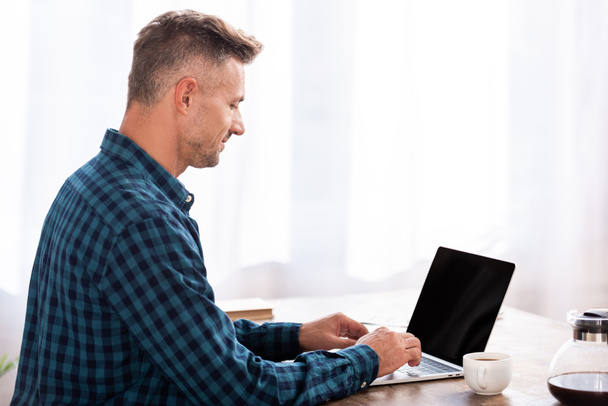 side view of smiling man in checkered shirt using laptop at home - Foto, Imagem