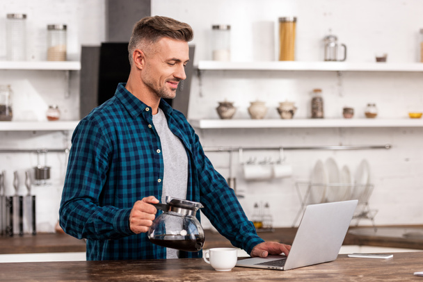 hymyilevä mies pitämällä kahvipannua ja käyttämällä kannettavaa kotona
 - Valokuva, kuva