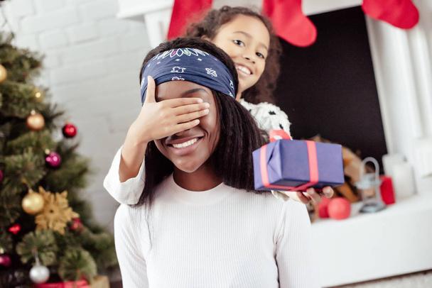 усміхнена афроамериканська дочка подарує мамі різдвяний подарунок і закриває очі на несподіванку вдома
 - Фото, зображення