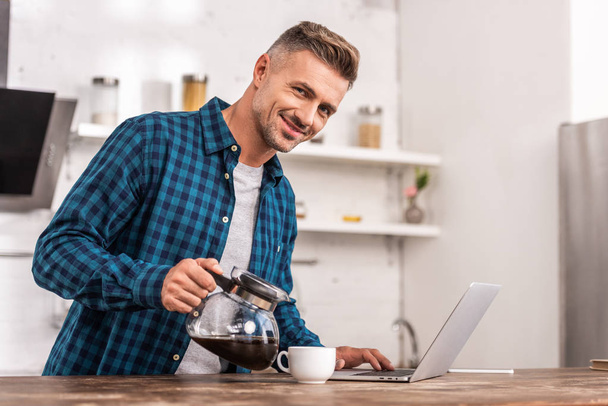 handsome smiling man pouring coffee and using laptop at home - Foto, Imagem