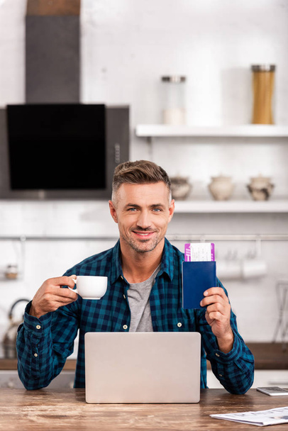 bel homme souriant tenant tasse de café et passeport avec carte d'embarquement tout en utilisant un ordinateur portable à la maison
  - Photo, image