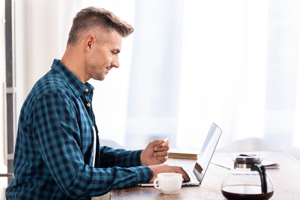 side view of smiling man holding credit card and using laptop at home - Foto, Imagem