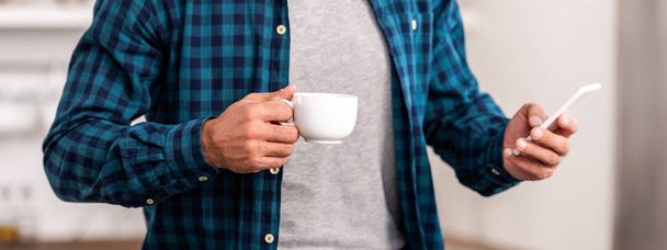 section médiane de l'homme en chemise à carreaux tenant tasse de café et en utilisant smartphone à la maison
  - Photo, image