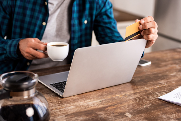 Nahaufnahme eines Mannes, der eine Tasse Kaffee und Kreditkarte in der Hand hält, während er zu Hause seinen Laptop benutzt - Foto, Bild