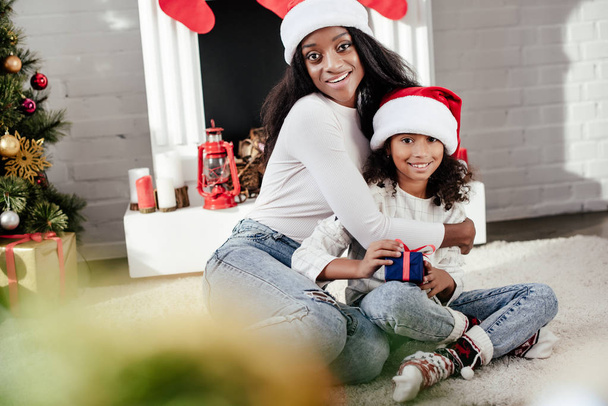smiling african american woman hugging little daughter with christmas gift in decorated room at home - Photo, Image