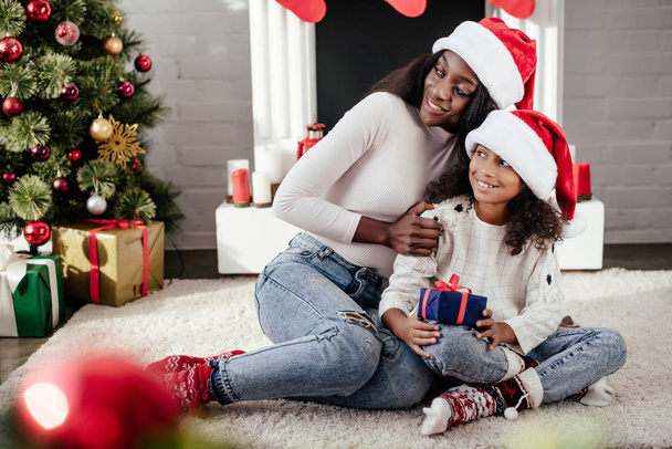 souriant afro-américaine femme étreignant petite fille avec cadeau de Noël dans la chambre décorée à la maison
 - Photo, image