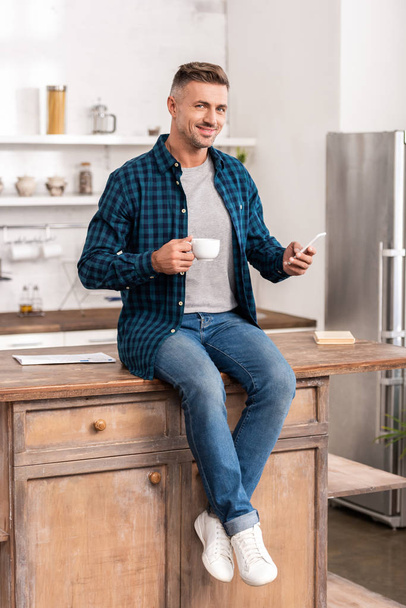 handsome smiling man holding cup of coffee and using smartphone while sitting on kitchen table - Φωτογραφία, εικόνα