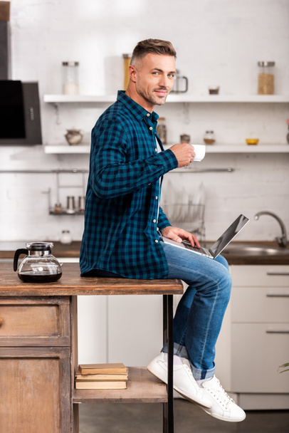 handsome adult man holding cup of coffee and smiling at camera while using laptop at home  - Fotografie, Obrázek