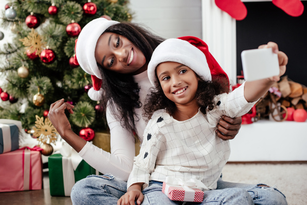african american family in santa claus hats taking selfie on smartphone in decorated room at home, christmas holiday concept - Photo, Image