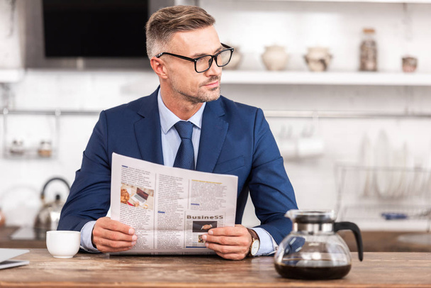handsome businessman in eyeglasses holding newspaper and looking away at home - Photo, Image